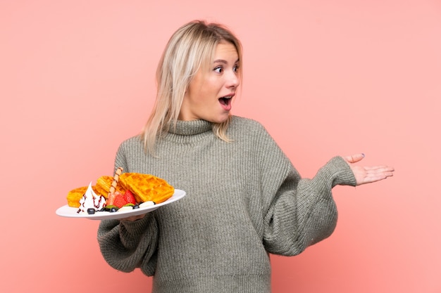 Jeune femme blonde tenant des gaufres sur un mur rose isolé avec une expression faciale surprise