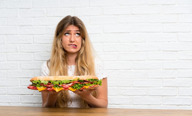 Jeune femme blonde tenant un gros sandwich ayant des doutes