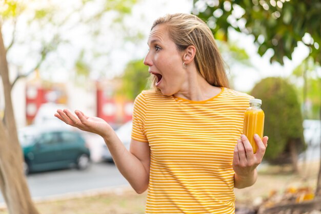 Photo une jeune femme blonde tenant un jus d'orange à l'extérieur avec une expression faciale de surprise