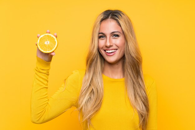 Jeune femme blonde tenant une orange sur un mur jaune