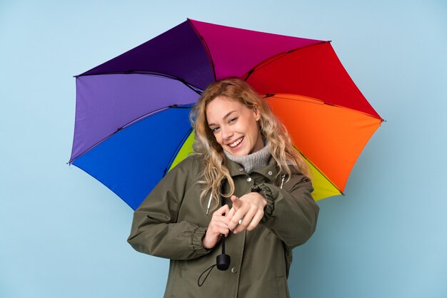 Jeune femme blonde tenant un parapluie isolé sur le mur bleu pointe le doigt vers vous avec une expression confiante