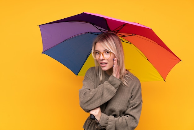 Jeune femme blonde tenant un parapluie sur mur jaune isolé chuchoter quelque chose