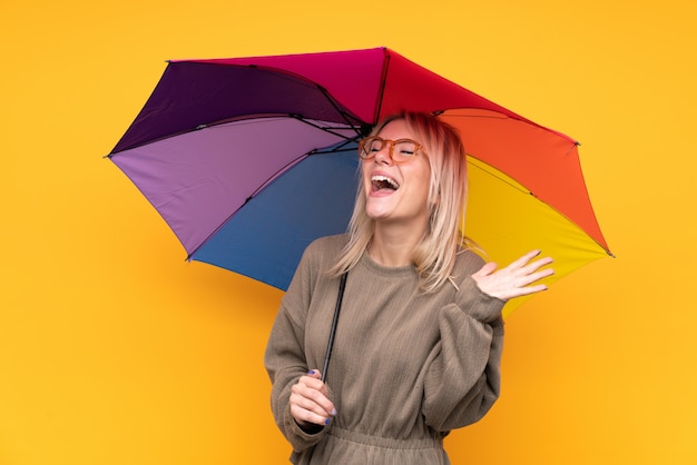 Jeune femme blonde tenant un parapluie sur un mur jaune isolé avec une expression faciale surprise