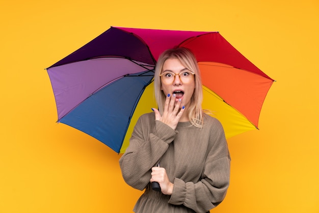 Jeune femme blonde tenant un parapluie sur un mur jaune isolé avec une expression faciale surprise