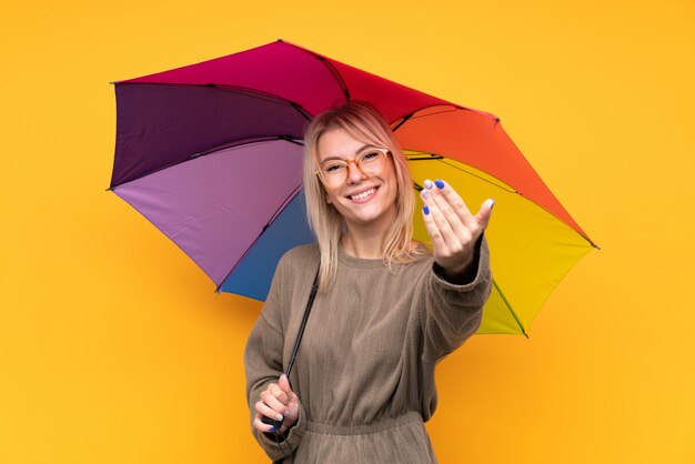 Jeune femme blonde tenant un parapluie sur un mur jaune isolé invitant à venir