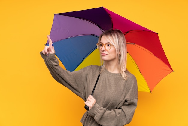 Jeune femme blonde tenant un parapluie sur un mur jaune isolé touchant sur un écran transparent