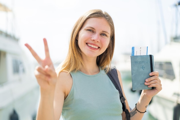 Photo jeune femme blonde tenant un passeport à l'extérieur souriant et montrant le signe de la victoire