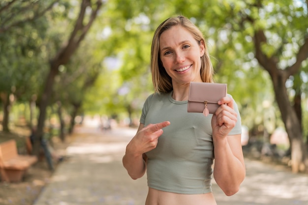 Une jeune femme blonde tenant un portefeuille à l'extérieur et le pointant