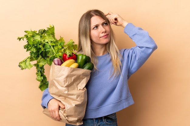 Jeune femme blonde tenant un sac plein de légumes sur un mur isolé ayant des doutes et avec une expression de visage confuse