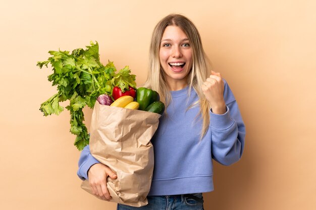 Jeune femme blonde tenant un sac plein de légumes sur un mur isolé célébrant une victoire