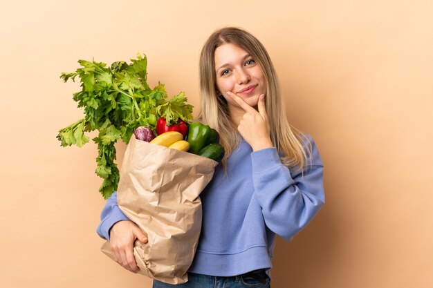 Jeune femme blonde tenant un sac plein de légumes sur rire isolé