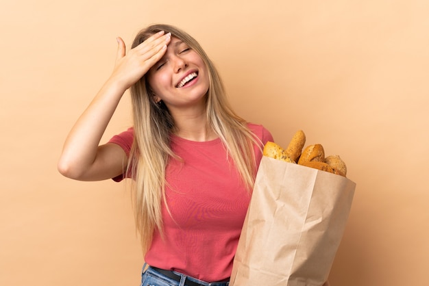 Jeune femme blonde tenant un sac plein de pains isolé sur un mur beige en riant