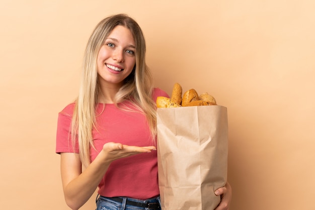 Jeune femme blonde tenant un sac plein de pains isolé sur un mur beige tendant les mains sur le côté pour inviter à venir