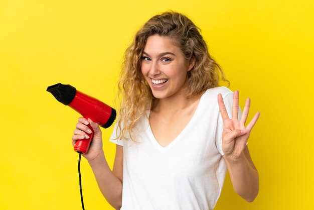 Jeune femme blonde tenant un sèche-cheveux isolé sur fond jaune heureux et comptant quatre avec les doigts