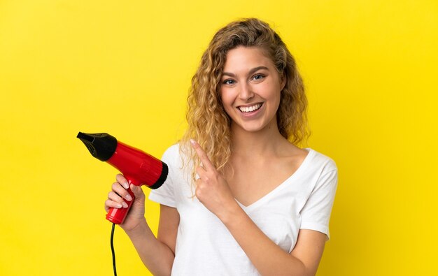 Jeune femme blonde tenant un sèche-cheveux isolé sur fond jaune pointant vers le côté pour présenter un produit
