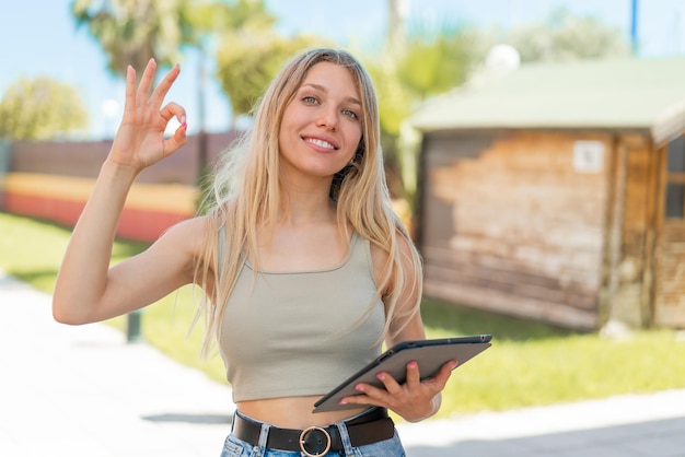 Jeune femme blonde tenant une tablette à l'extérieur montrant le signe ok avec les doigts