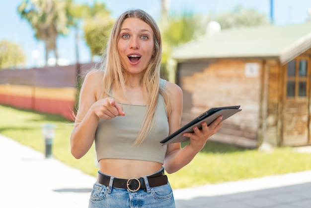 Jeune femme blonde tenant une tablette à l'extérieur surpris et pointant vers l'avant