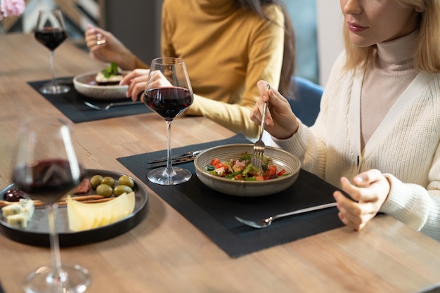 Jeune femme blonde en tenue décontractée élégante assise près d'une table servie contre son amie et ayant une salade de légumes, du vin rouge, du fromage et des olives