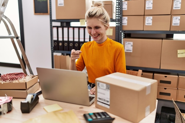 Jeune femme blonde travaillant dans une petite entreprise de commerce électronique à l'aide d'un ordinateur portable souriant heureux pointant avec la main et le doigt
