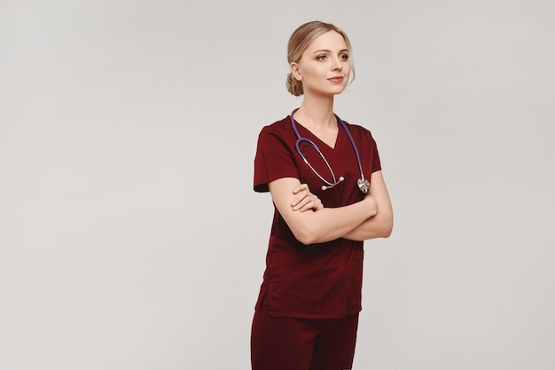 Jeune femme blonde en uniforme médical avec stéthoscope posant avec les mains jointes