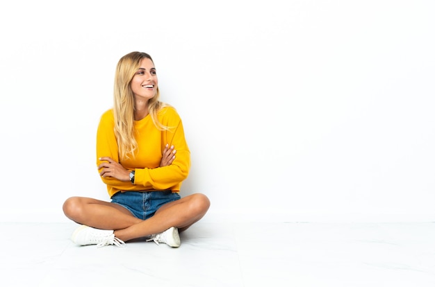 Jeune femme blonde uruguayenne assise sur le sol isolé sur un mur blanc heureux et souriant