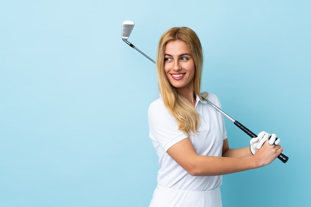 Jeune femme blonde uruguayenne sur mur bleu isolé jouant au golf