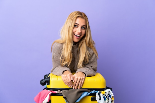 Jeune femme blonde uruguayenne avec une valise pleine de vêtements sur mur violet isolé