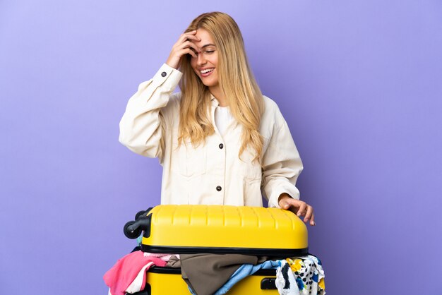 Jeune femme blonde uruguayenne avec une valise pleine de vêtements sur mur violet rire