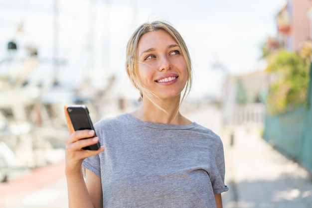 Jeune femme blonde utilisant un téléphone portable à l'extérieur en levant les yeux en souriant