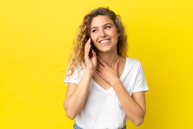 Jeune femme blonde utilisant un téléphone portable isolé sur fond jaune en levant tout en souriant