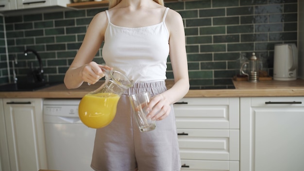 Une jeune femme blonde verse du jus d'orange dans un verre et le boit. Une femme assez mince boit du jus d'orange juteux dans la cuisine. La caméra se déplace de bas en haut, 4K UHD.