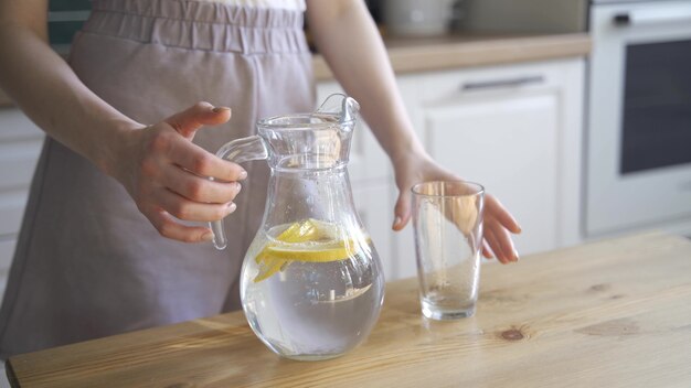 Une jeune femme blonde verse de l'eau citronnée d'une carafe dans un verre. Eau rafraîchissante avec des tranches de citron. 4K UHD