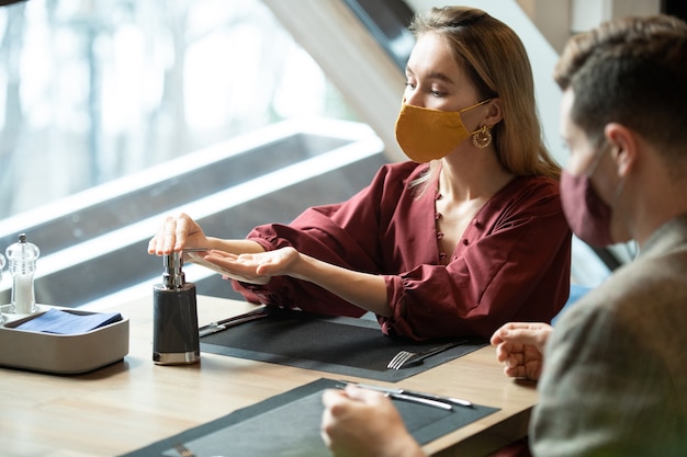 Jeune femme blonde en vêtements de sport intelligents et masque de protection en appuyant sur le distributeur de pot en plastique avec désinfectant pour les mains avant de dîner au café