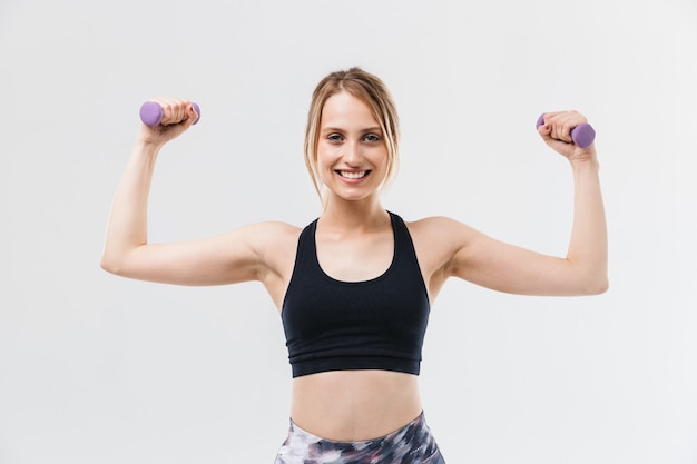 jeune femme blonde vêtue de vêtements de sport faisant de l'exercice et faisant des exercices avec des haltères pendant la remise en forme dans une salle de sport isolée sur un mur blanc