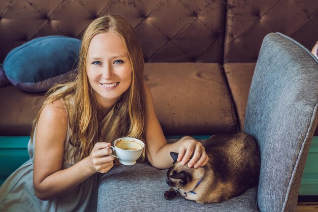 La jeune femme boit du café et caresse le chat Dans le contexte du canapé rayé par les chats