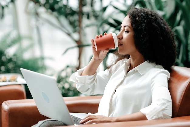 La jeune femme boit du café tout en travaillant sur un ordinateur portable