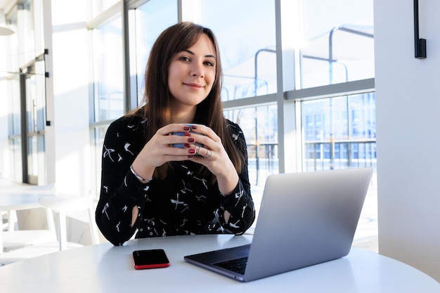 Une jeune femme boit du café et travaille sur un ordinateur portable dans un caféModèle élégant regarde dans l'appareil photo