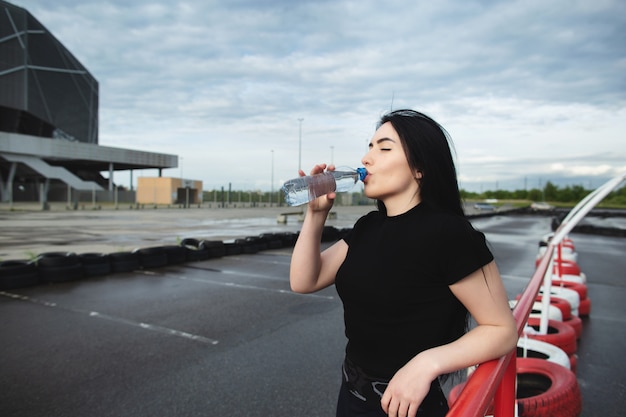 Jeune femme boit de l'eau d'une bouteille après une course, en appuyant sa main sur la barre horizontale. Femme athlétique s'entraîne à l'extérieur près du stade. Soins de santé, concept sportif