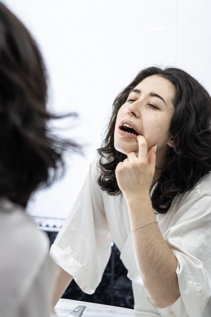 Jeune femme avec la bouche ouverte vérifiant les dents dans le miroir dans la salle de bain à la maison femme brune regardant les dents de la bouche dans le meroir de la salle de bains avant soin des dents concept de beauté et de santé