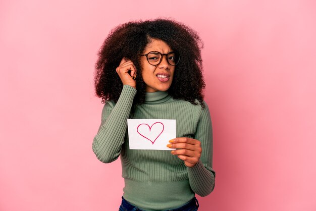 Jeune femme bouclée afro-américaine tenant un symbole du cœur sur une pancarte couvrant les oreilles avec les mains.