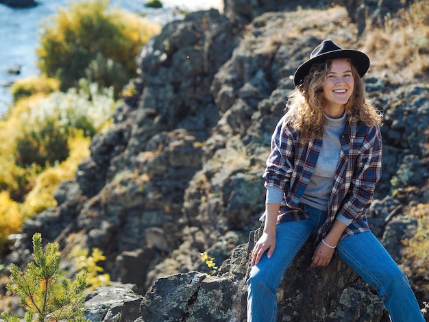 Photo jeune femme bouclée en chapeau et t-shirt à carreaux souriant sur la colline de la montagne avec vue sur la rivière automne