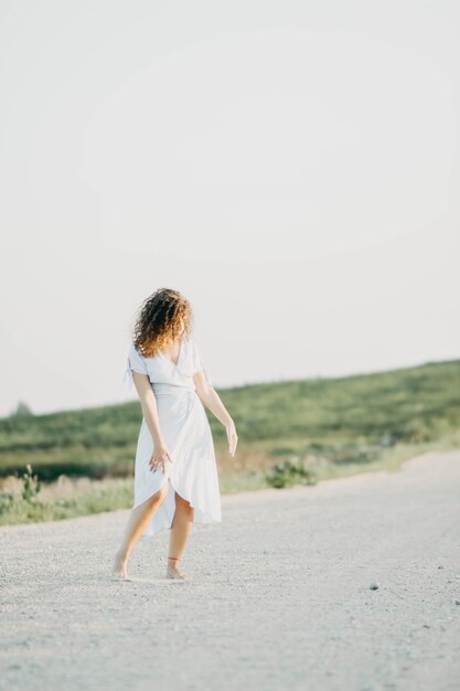 jeune femme bouclée dansant romantiquement dans une robe bleue sur une route sablonneuse au coucher du soleil