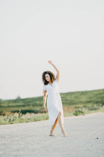 jeune femme bouclée dansant romantiquement dans une robe bleue sur une route sablonneuse au coucher du soleil