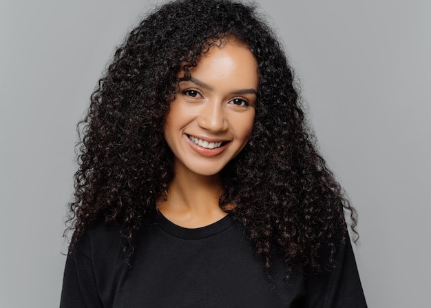 Une jeune femme bouclée optimiste avec un sourire doux regarde positivement la caméra porte des vêtements noirs décontractés isolés sur fond gris écoute une histoire intéressante ou une anecdote amusante Concept d'émotions