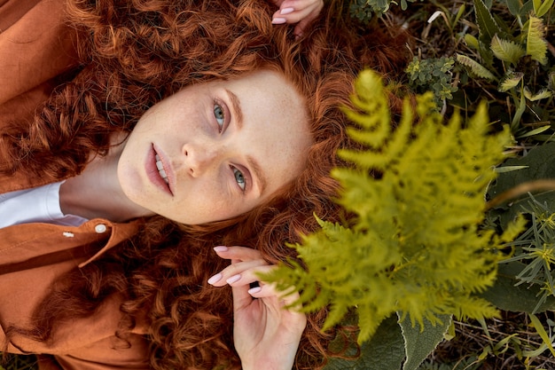 Jeune femme bouclée rousse d'apparence caucasienne allongée sur l'herbe en été posant en regardant la caméra...