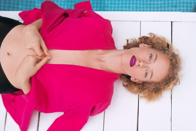 Jeune femme bouclée rousse portant une veste fuchsia au bord de la piscine