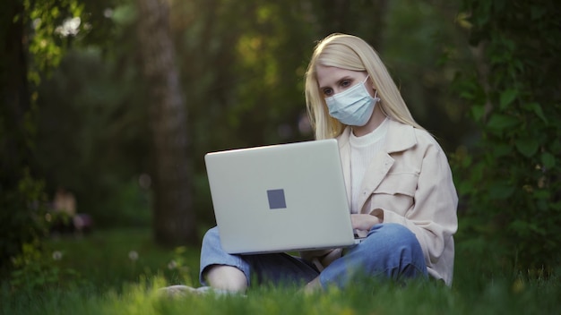 Jeune femme avec des boucles dans un masque médical travaille à l'aide d'un ordinateur portable dans le parc. Travail à distance pendant le confinement. Travail en extérieur. 4K UHD