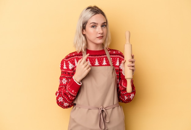 Jeune femme boulangère caucasienne faisant des biscuits pour Noël isolée sur fond jaune pointant le doigt vers vous comme si vous vous invitiez à vous rapprocher.