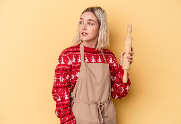 Jeune femme boulangère caucasienne faisant des cookies pour Noël isolée sur fond jaune regarde de côté souriante, gaie et agréable.