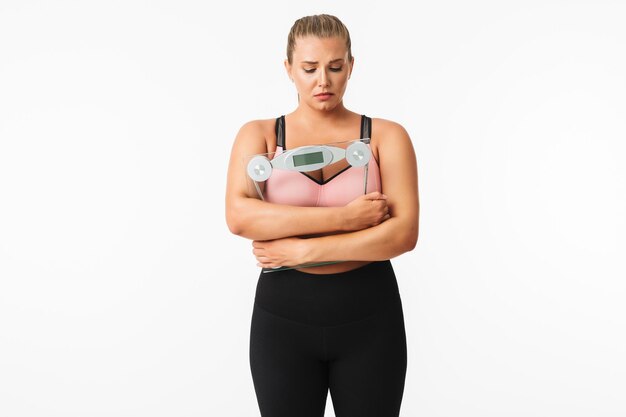 Jeune femme bouleversée avec un excès de poids en haut sportif tenant des balances dans les mains sur fond blanc isolé